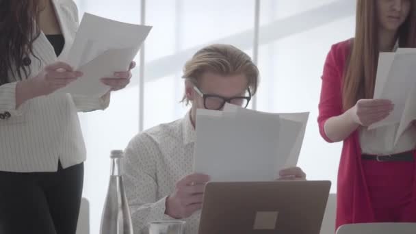 Portrait tired man sitting in a comfortable office with netbook at the table examining documents which female colleagues gives to him one by one. Two girls in formal wear standing on either side of — Stock Video