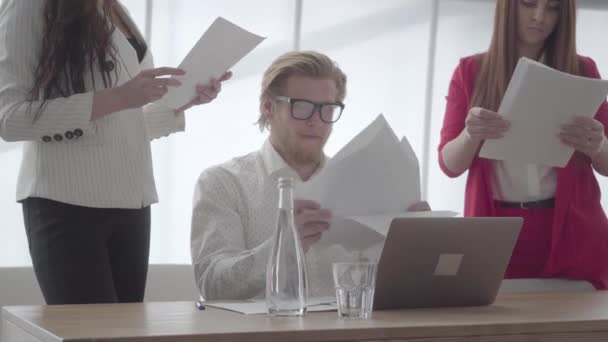 2人の女性の同僚が一人ずつ彼に与える書類を収集し、その後、紙のスタックを投げるテーブルでネットブックを持って現代のオフィスに座っている肖像画神経質な男。上司は疲れていて — ストック動画