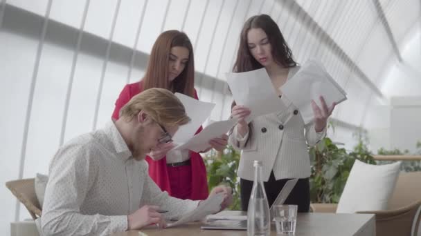 Portret vermoeide man zittend in een comfortabel kantoor met netbook aan de tafel het bestuderen van documenten die vrouwelijke collega's hem één voor één geven. Twee meisjes in formele slijtage staande aan beide zijden van — Stockvideo