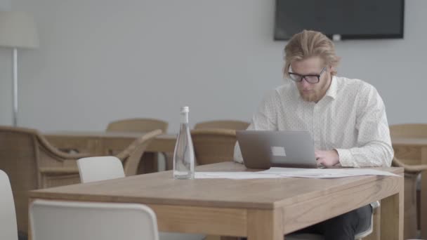 Πορτρέτο επιτυχημένος ξανθός άνθρωπος με γυαλιά που κάθονται στο τραπέζι σε ένα ελαφρύ άνετο γραφείο μπροστά από netbook. Ωραίος επιχειρηματίας που εμπλέκεται στη διαδικασία εργασίας. Η κάμερα μετακινείται δεξιά — Αρχείο Βίντεο