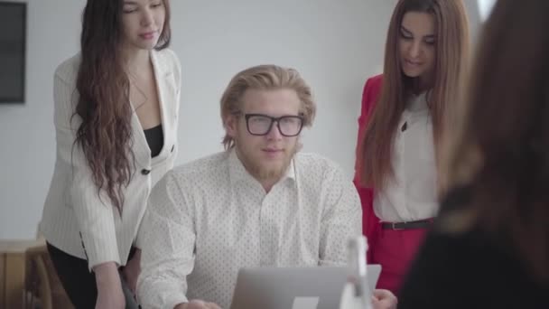 Blonder Mann mit Brille sitzt in einem hellen, gemütlichen Büro mit Netbook am Tisch. Zwei Mädchen in offizieller Kleidung stehen neben ihm und eine sitzt vor ihm und gibt dem Chef Dokumente. Arbeit — Stockvideo