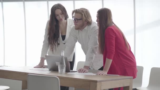 Hombre de negocios guapo en gafas dobladas sobre netbook en una oficina cómoda luz que muestra información a colegas femeninos. Dos mujeres en ropa formal de pie a cada lado del jefe — Vídeo de stock