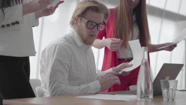 Porträt eines erfolgreichen blonden Mannes mit Brille, der in einem hellen, gemütlichen Büro mit Netbook am Tisch sitzt und Dokumente signiert, die ihm Kollegen mitgebracht haben. zwei Mädchen in formeller Kleidung stehen auf einem der beiden — Stockvideo