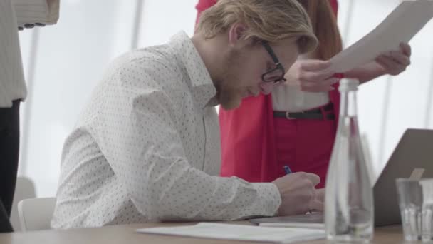 Portrét úspěšného světlovlasého muže v brýlích sedícího v lehké pohodlné kanceláři s netbookem u stolu podepisující dokumenty, které mu kolegové přinesli. Dvě dívky ve formálním oblečení, které stojí na — Stock video