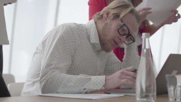 Porträt eines erfolgreichen blonden Mannes mit Brille, der in einem hellen, gemütlichen Büro mit Netbook am Tisch sitzt und Dokumente signiert, die ihm Kollegen mitgebracht haben. zwei Mädchen in formeller Kleidung stehen auf einem der beiden — Stockvideo