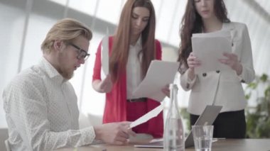 Portre sinir adam masada netbook ile modern ofiste oturan iki kadın meslektaşları ona birer birer verir belgeleri toplama, sonra kağıt yığını atmak.