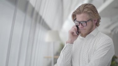Yakışıklı üzgün adam gözlüklü hafif rahat bir ofiste cep telefonu ile konuşurken duruyor. Yakışıklı bir işadamı işine dahil oldu. İşadamı cep telefonuyla önemli bir konuşma yaptı