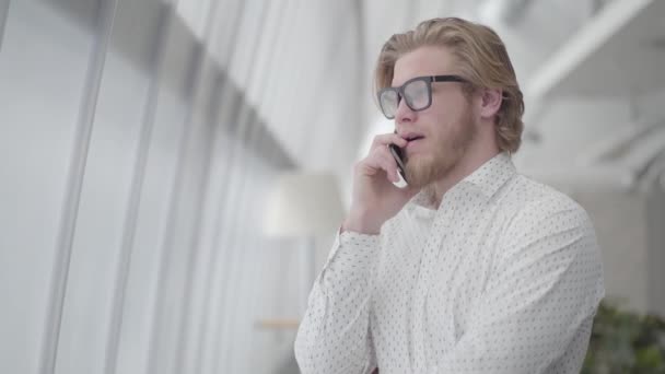 Ξανθός νευρικός άνθρωπος με γυαλιά στέκεται σε ένα ελαφρύ άνετο γραφείο μιλάει με το κινητό. Ωραίος επιχειρηματίας που εμπλέκεται στη δουλειά του. Ο επιχειρηματίας έχει σημαντική συζήτηση μέσω κινητού τηλεφώνου — Αρχείο Βίντεο
