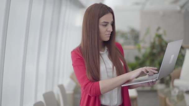 Młoda kobieta w czerwonym garniturze pisząca na swoim gadżecie w nowoczesnym biurze. Dziewczyna zamyka swój netbook i uśmiecha się. Pracownik biurowy ma dobre wieści. Miejsce pracy. Powierzchnia biurowa. — Wideo stockowe