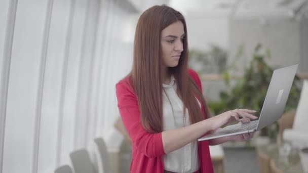 Mulher nova no terno vermelho que datilografa em seu gadget no escritório claro moderno. A menina fecha o netbook e sorri. O funcionário do escritório tem boas notícias. Local de trabalho. Espaço de escritório . — Vídeo de Stock