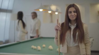 Resmi giyim bilardo işareti tutan zarif kadın portresi ve iki bulanık erkek ve kadın figürleri önünde kamera bakıyor. Bayan arkadaşlarıyla bilardo oynuyor.
