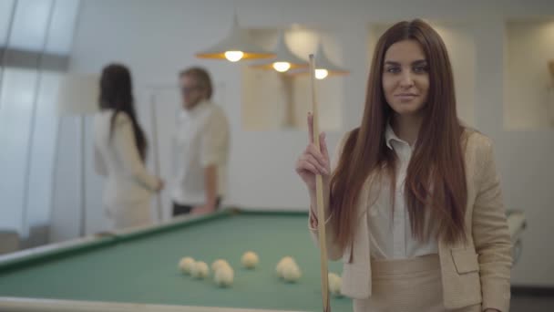 Retrato de una linda mujer joven en ropa formal sosteniendo la señal de billar y mirando en cámara delante de dos figuras masculinas y femeninas borrosas. La señora juega al billar con sus amigos — Vídeo de stock