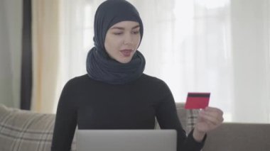 Modern Müslüman elbiseli, güzel başlıklı, gülümseyen doğulu genç bir kadın dizüstü bilgisayarla internet üzerinden ödeme yapmak için kredi kartı kullanıyor. Türbanlı genç Asyalı Müslüman kadın internet dükkanından bir şeyler alıyor.