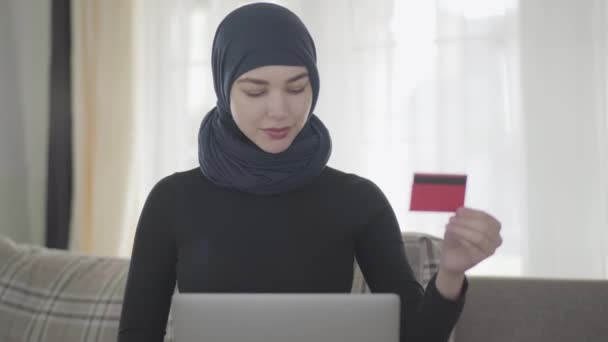 Retrato joven mujer musulmana sonriente utilizando una tarjeta de crédito para pagar en línea utilizando el ordenador portátil. Chica joven en pañuelo para la cabeza comprar artículos en la tienda de Internet . — Vídeo de stock