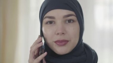 Modern Müslüman giysileri ve güzel siyah başlık cep telefonu ile konuşurken yakın genç bir güzel doğu kadın Portresi. Başörtülü genç Asyalı Müslüman kadın kameraya bakıyor
