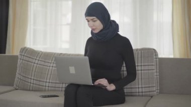 Balkon elbiseli, güzel başlıklı, evinde kanepede laptopuyla çalışan başarılı, kendine güvenen genç bir kız. Türbanlı genç Asyalı Müslüman kadın internette sörf yapıyor.