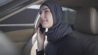 Modern Müslüman kıyafetleri içinde gülümseyen ve arabada cep telefonuyla konuşan güzel başlıklı genç bir kadın. Türbanlı Asyalı kadın otomobil kullanıyor.