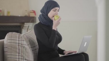 Genç Müslüman kadın laptopun başında çalışıyor ya da sohbet ediyor ve geleneksel başörtüsü takarak elma ısırıyor.
