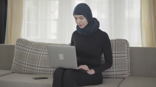 Portrétní mladý úsměv úspěšné sebevědomé dívky v balcového oblečení a nádherný Heš pracující s přenosným notebookem, který sedí na pohovce doma. Mladá Asijská muslimka v šátku na internetu — Stock video