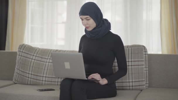 Jong lachend succesvol zelfverzekerd meisje in kale kleren en mooie hoofdtooi werkend met haar laptop zittend op de bank thuis. Jonge aziatische moslim vrouw in hoofddoek surfen op het web — Stockvideo