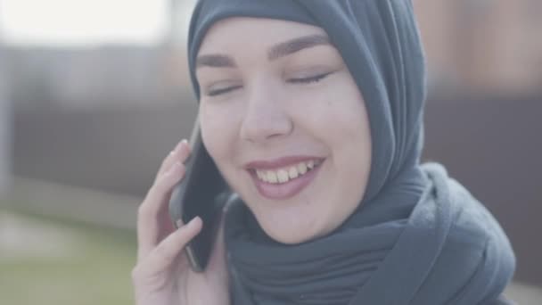 Närbild porträtt av en ung vacker muslimsk kvinna i svart huvudbonat pratar med mobiltelefon närbild. Söt asiatisk tjej i huvudscarf tittar i kameran. — Stockvideo