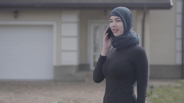 Porträtt av en ung vacker muslimsk kvinna i svart huvudbonat pratar med mobiltelefon utomhus. Söt asiatisk tjej i huvudscarf tittar i kameran. — Stockvideo