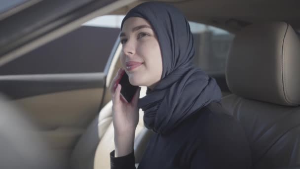 Mladá usměvavá východní žena v moderních muslimských šatech a s krásnou čelenkou u mobilu sedící v autě. Asijské žena v headscarf řízení automobil — Stock video