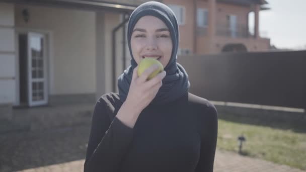 Retrato de mulher muçulmana jovem independente mordendo maçã verde suculenta usando lenço de cabeça tradicional no fundo da casa — Vídeo de Stock