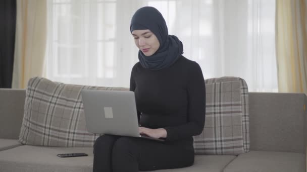 Succesvol zelfverzekerd meisje in Balck kleren en mooie hoofdtooi werken met haar laptop zittend op de Bank thuis. Jonge Aziatische moslim vrouw in hoofddoek surfen op het web — Stockvideo