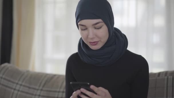 Ung vacker östlig kvinna i svarta kläder och vacker huvudbonad ringer via mobiltelefon. Ung asiatisk muslimsk dam i huvudduk med gadget sitter på soffan hemma tittar på kameran — Stockvideo