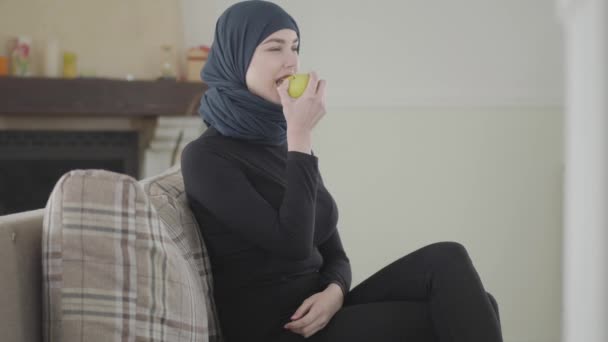 Красивая мусульманка смотрит телешоу и кусает яблоко в традиционном платке на фоне уютной гостиной — стоковое видео
