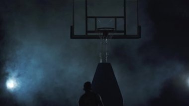 Kırmızı üniformalı genç basketbolcu topu sepetin içine atar. Siyah arka planda duman bulutunun içinde. Profesyonel basketbol oyuncusu iş başında. Spor anlayışı