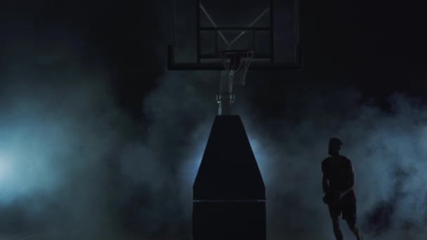 Gezonde jonge man speelt basketbal in zwarte achtergrond met wolken van rook. Het dragen van rode shirt en zwarte shorts. — Stockvideo