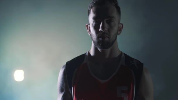 Pohledný muž v červené uniformě hází míč a otáčí se na prstech a dívá se do kamery před reflektoru oblaku kouře v pozadí. Profesionální hráč basketbalového hry — Stock video