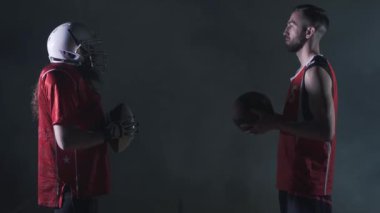 Farklı oyunların iki erkek spor oyuncuları karanlık arka planda birbirlerinin önünde duruyor. Amerikalı futbolcu ve basketbolcu birbirlerine top tutan bakmak. Yan görünüm