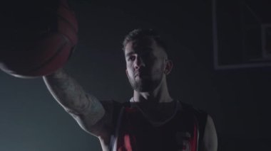 Yakın portre kırmızı üniformalı yakışıklı kendine güvenen adam karanlıkta uzanmış elinde bir top tutarak. Profesyonel basketbol oyunu oyuncu bir top ile oynuyor. Arka plandaki sepet