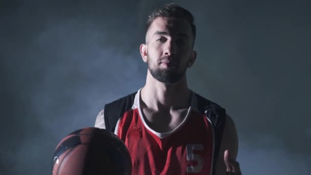 Un bel homme confiant en uniforme rouge tenant une balle dans la main regardant la caméra dans le fond sombre dans un nuage de fumée. Joueur de basket professionnel, vue de face — Video