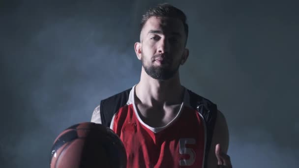 Kırmızı üniformalı yakışıklı kendine güvenen adam elinde bir top tutarak duman bulutu içinde karanlık arka planda kameraya bakıyor. Profesyonel basketbol oyuncusu, ön görünüm — Stok video