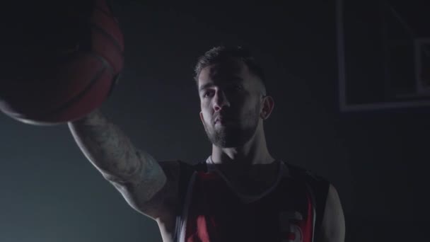 Yakın portre kırmızı üniformalı yakışıklı kendine güvenen adam karanlıkta uzanmış elinde bir top tutarak. Profesyonel basketbol oyunu oyuncu bir top ile oynuyor. Arka plandaki sepet — Stok video