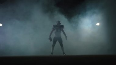 Amerikan futbol sporcusu futbol kask elinde topu ile duman bulutu içinde spot ile siyah arka plan üzerinde sahada duran