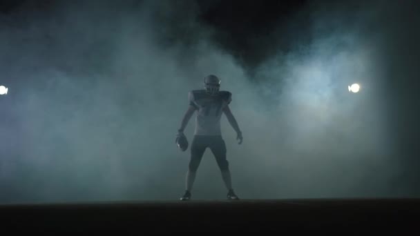 Hráč amerického fotbalového sportovce v fotbalové helmě stojící na poli na černém pozadí se světlem v oblaku kouře s koulí v rukou — Stock video