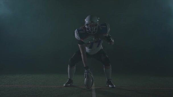 Американский футболист в футбольном шлеме стоит на линии на поле, держа мяч, и готовится бежать на черном фоне с мигающим светом. Вид спереди — стоковое видео