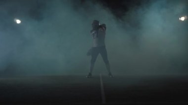 Amerikan futbol sporcusu futbolcu portresi elinde topu ile duman bulutu siyah arka plan üzerinde sahada duran futbol kask. Top kaldıran adam çağıran jestler yapar