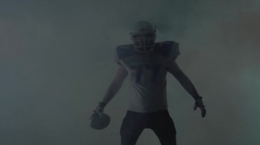 Portre amerikan futbol sporcusu futbol kask elinde topu ile duman bulutu siyah arka plan üzerinde sahada duran. Top kaldıran adam çağıran jestler yapar