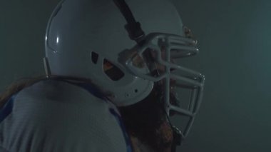 Kasklı sakallı Amerikan futbolcusunun yakın portresi koşmaya başlar ve karanlık arka planda bir çizgi çizer. Erkeklik kavramı, spor, zafer. Yan görünüm