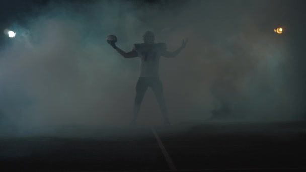 Desportista de futebol americano jogador no capacete de futebol em pé no campo em fundo preto em uma nuvem de fumaça com a bola nas mãos. O homem levantando bola faz gestos invocando — Vídeo de Stock