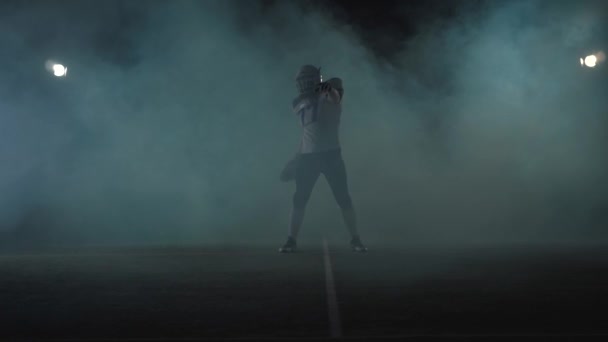 Porträt eines amerikanischen Fußballspielers mit Fußballhelm, der auf dem Spielfeld vor schwarzem Hintergrund in einer Rauchwolke steht und den Ball in der Hand hält. Der Mann, der den Ball hebt, macht anrufende Gesten — Stockvideo