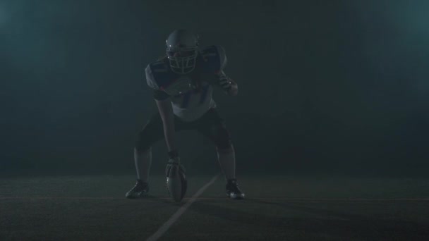 フットボールのヘルメットを着用したアメリカンフットボールのスポーツマン選手は、彼のボールを保持し、点滅する光で黒い背景に実行する準備をフィールド上のラインに立っています。正面図 — ストック動画