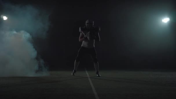 American football sportsman player in football helmet standing on the field on black background in a cloud of smoke with the ball in hands. The man raising ball makes invoking gestures — Stock Video