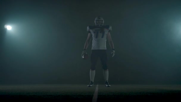 Portret American Football sportman speler in voetbal helm staande op het veld op zwarte achtergrond in een wolk van rook met de bal in handen. De man Raising Ball maakt het maken van bedreigend — Stockvideo